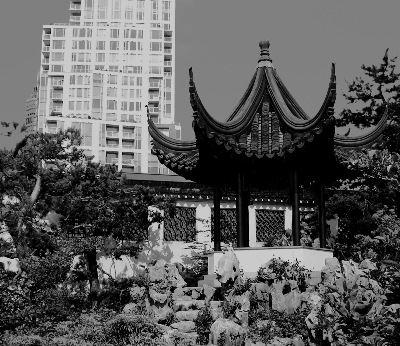 A black & white image of the Dr. Sun Yat-Sen Classical Chinese Garden in Vancouver, BC, Canada; showing the Chinese lake pavilion, with modern sky-scrapper building architecture in the background.